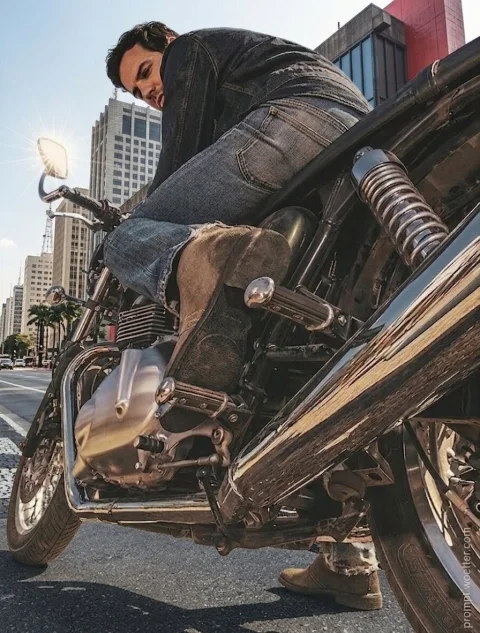 Fotografia em ângulo baixo (contra-plongée) de um homem montado em uma motocicleta clássica estacionada em uma via urbana. O primeiro plano destaca em detalhes o escapamento cromado longo, as molas da suspensão e o motor metálico. O motociclista veste calça jeans, jaqueta escura e botas de camurça marrom. Um forte reflexo de sol em formato de estrela brilha no retrovisor, com arranha-céus ao fundo.