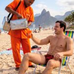 Fotografia em plano inteiro em uma praia ensolarada no Rio de Janeiro, com o morro Dois Irmãos ao fundo. Um homem sorridente, sem camisa e de sunga vermelha estampada, está sentado em uma cadeira de praia listrada e colorida, estendendo um copo laranja para receber a bebida. À esquerda, um vendedor ambulante negro veste o clássico uniforme laranja dos vendedores de mate cariocas e carrega galões de metal a tiracolo, servindo o chá mate no copo do homem. No céu azul com poucas nuvens, um pequeno avião monomotor voa puxando uma faixa vermelha onde se lê em letras brancas: "DIA INTERNACIONAL DO CHÁ".
